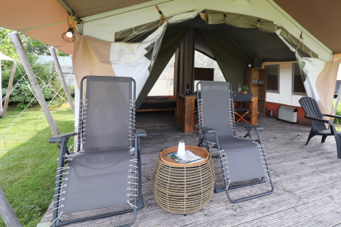 Tente safari avec terrasse en bois, deux chaises longues, une table ronde et intérieur aménagé au fond.
