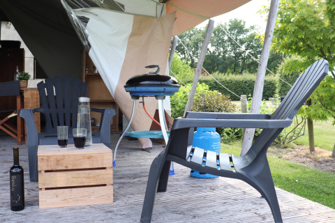 Terrasse d'une tente safari avec chaises en plastique, table en bois, bouteille de vin et barbecue.
