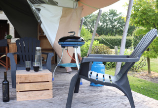 Terrasse d'une tente safari avec chaises en plastique, table en bois, bouteille de vin et barbecue.
