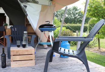 Safari tent patio with plastic chairs, a wooden table, a wine bottle, a carafe, and a barbecue grill.