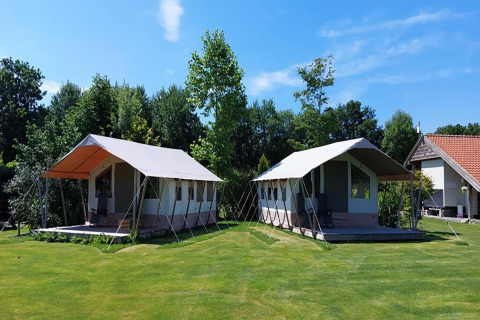 Deux tentes safari installées sur une pelouse verte, entourées d’arbres et sous un ciel bleu clair.