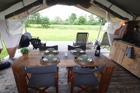 Intérieur d’une tente safari avec table en bois, vaisselle, coin cuisine et vue sur une prairie verte.