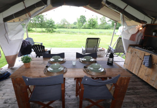 Intérieur d’une tente safari avec table en bois, vaisselle, coin cuisine et vue sur une prairie verte.