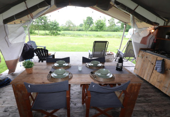 Safari tent interior with wooden table, set dishes, a small kitchen, and sunny green field view outside.