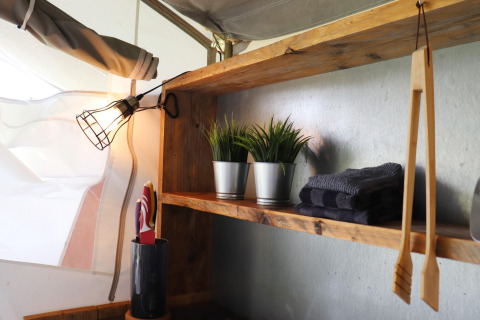 Étagère en bois dans une tente safari avec lampe, serviettes grises, plantes artificielles et ustensiles.
