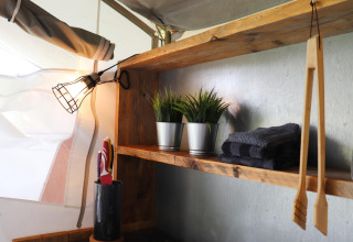 Étagère en bois dans une tente safari avec lampe, serviettes grises, plantes artificielles et ustensiles.