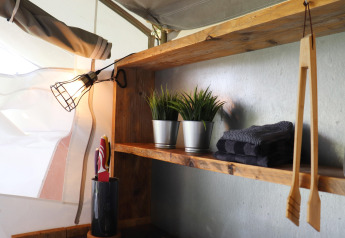 Wooden shelf in a safari tent featuring an exposed lamp, towels, artificial plants, and utensils.
