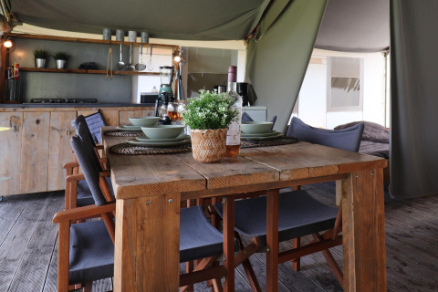 Coin repas dans une tente safari avec table en bois rustique, chaises, cuisine et vaisselle dressée.
