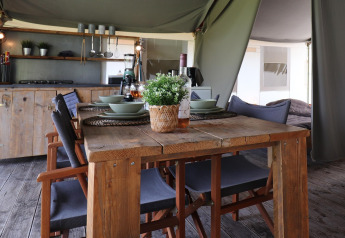 Dining area inside a safari tent, featuring a rustic wooden table, chairs, kitchen, and table setting.