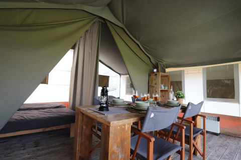 Intérieur d'une tente safari avec table à manger en bois, quatre chaises et lit visible en arrière-plan.