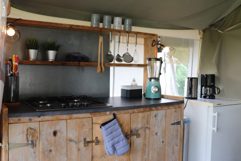 Coin cuisine dans une tente safari avec armoire en bois, réchaud, frigo, blender, cafetière et ustensiles.
