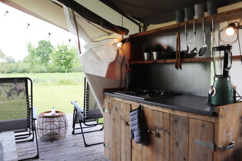 Safari tent kitchen with wooden cabinets, gas cooktop, blender, patio chairs, and view of grass.