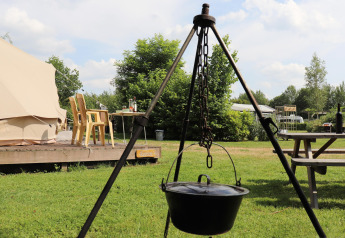Photo d’un hébergement glamping : tente, table, chaises et marmite suspendue au feu en pleine nature.