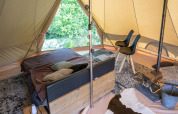 Innenaufnahme eines Glamping-Zeltes mit Bett, Stühlen, Teppichen und Holzofen in grüner Umgebung.