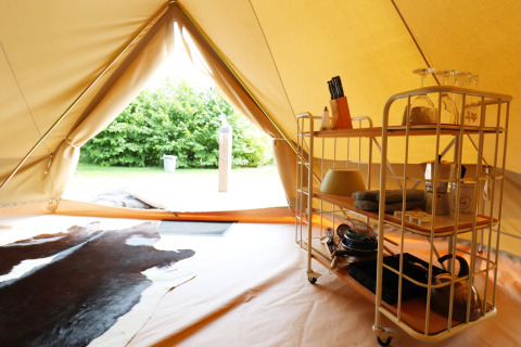 Dentro una tenda glamping con carrello cucina, stoviglie, tappeto in pelle e vista sull’esterno verde.