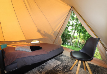 Intérieur d’une tente de glamping avec un lit simple, une chaise et une vue sur des arbres verts à l'extérieur.