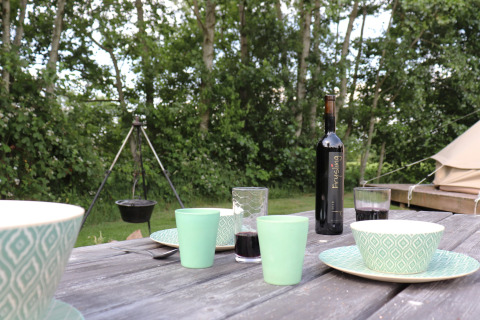 Buitentafel met bekers, kommen en wijnfles, glamping tent en hangende pot op de achtergrond.