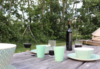Udendørs bord med kopper, skåle og vinflaske, glamping telt og gryde over bål i baggrunden.