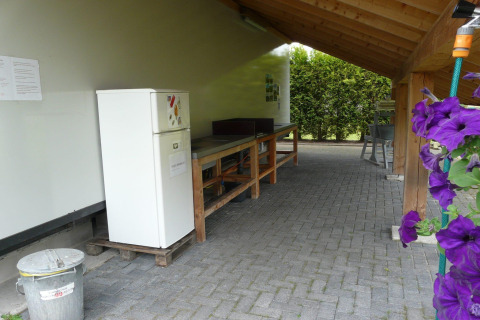 Udendørs glampingkøkken med et køleskab, langbord og blomster under en trætagskonstruktion.