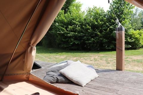 Uitzicht vanuit een glamping-tent met kussens op een houten terras en groene struiken op de achtergrond.