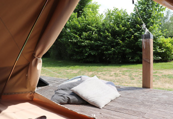 Vue d'une tente de glamping avec des coussins sur une terrasse en bois et des buissons verts au fond.
