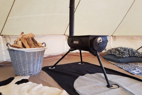 Cozy glamping tent interior featuring a wood-burning stove, firewood basket, and comfortable bedding.
