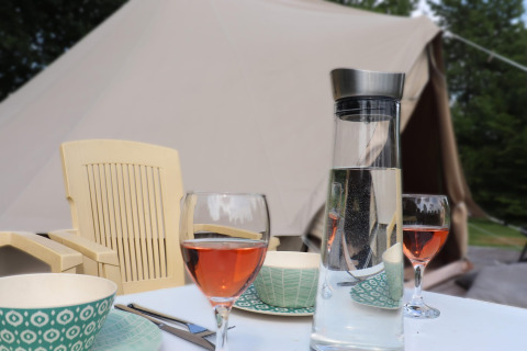 Mesa de glamping al aire libre con copas de vino rosado, jarra de agua, platos y tienda de campaña detrás.