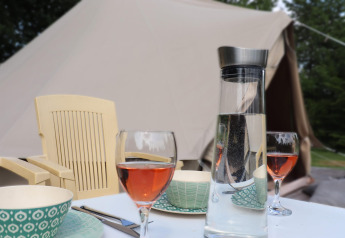 Moment de glamping avec table dressée, verres de vin rosé, carafe d'eau et tente en toile à l’arrière-plan.