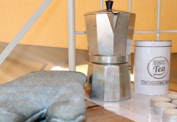 A moka pot, tea tin, and oven mitt on a table inside a cozy glamping tent with warm lighting.