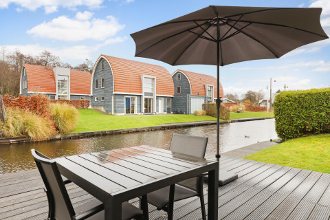 Terraza junto al canal en el Water park De Bloemert 6, Países Bajos, con modernas cabañas y sombrilla.