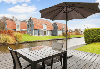 Terraza junto al canal en el Water park De Bloemert 6, Países Bajos, con modernas cabañas y sombrilla.