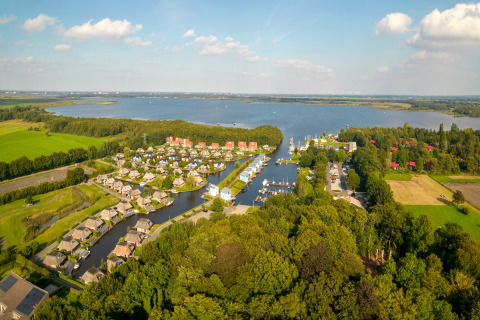 Luftaufnahme einer Lodge am See mit umliegenden Häusern, Wiesen und Wäldern bei Sonnenschein.