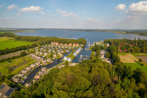Veduta aerea di un lodge sul lago circondato da case, campi verdi, boschi e acqua blu tranquilla.