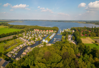 Luftaufnahme einer Lodge am See mit umliegenden Häusern, Wiesen und Wäldern bei Sonnenschein.