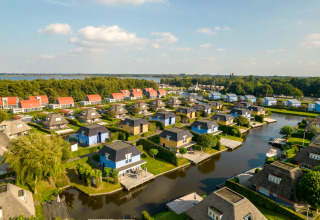Luftaufnahme einer Ferienanlage mit bunten Lodges, Kanälen und Booten bei einem See und Wald.