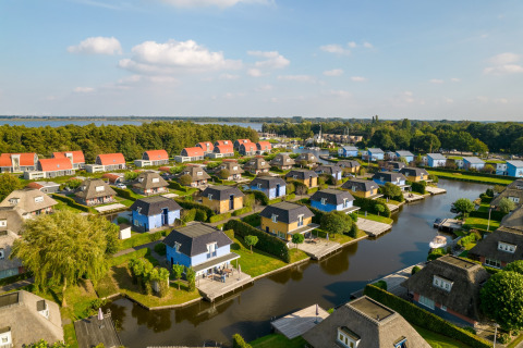 Luftaufnahme einer Ferienanlage mit bunten Lodges, Kanälen und Booten bei einem See und Wald.