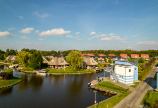 Vista panoramica di lodge lungo un canale con barche, alberi verdi e cielo blu in una giornata di sole.