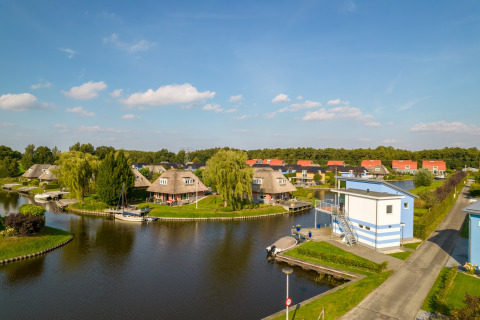 Panoramic view of lodges by a canal with boats, green trees, and blue skies on a sunny day.