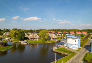 Panoramic view of lodges by a canal with boats, green trees, and blue skies on a sunny day.