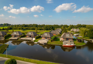 Luftfoto af idylliske sommerhuse ved en sø, omgivet af grønt landskab og blå himmel i baggrunden.