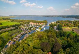 Vue aérienne d’un lodge au bord du lac entouré de champs verts, forêt et petites maisons au bord de l’eau.