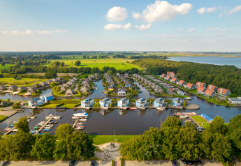 Luftaufnahme einer modernen Ferienanlage mit Hütten am See, Yachthafen und grüner Landschaft.