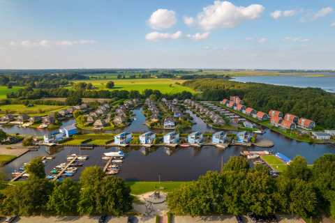 Luftaufnahme einer modernen Ferienanlage mit Hütten am See, Yachthafen und grüner Landschaft.