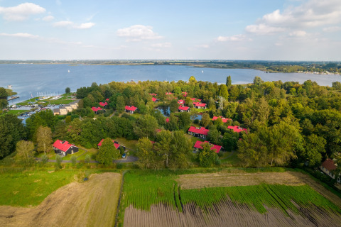Vue aérienne d’un lodge paisible au bord d’un lac, cabanes aux toits rouges, arbres et champs autour.