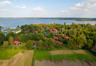 Vista aérea de una acogedora cabaña junto al lago, rodeada de bosque y campos, con techos rojos.