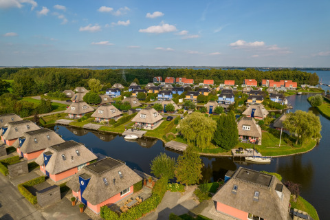Luftfoto af idylliske lodgehuse med stråtag ved en kanal med både, omgivet af frodigt grønt landskab.