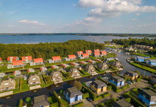 Vue aérienne de lodges pittoresques au bord d’un lac, toits rouges, pelouses vertes et forêt autour.