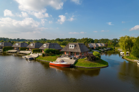 Foto von malerischen Lodges am Wasser mit Boot vor Anlegestelle, viel Grün und blauem Himmel.