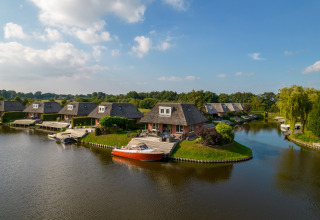 Photo de lodges au bord de l’eau avec toits de chaume, bateaux amarrés et verdure sous ciel bleu.