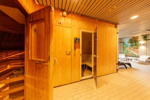 Ingang van een houten sauna in een ontspanningsruimte met ligstoelen en een muurschildering op de achtergrond.